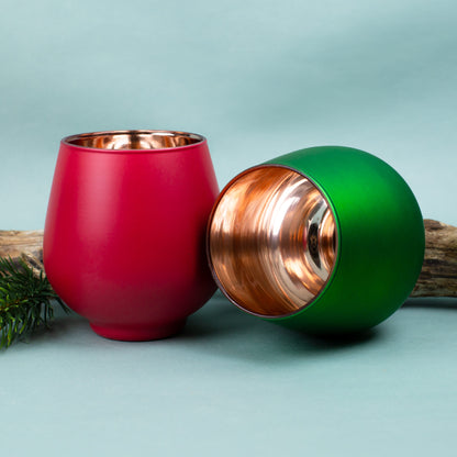 Rotes und grünes Windlicht mit kupferfarbener Innenfläche, dekorativ auf hellem Untergrund mit Tannenzweig platziert.