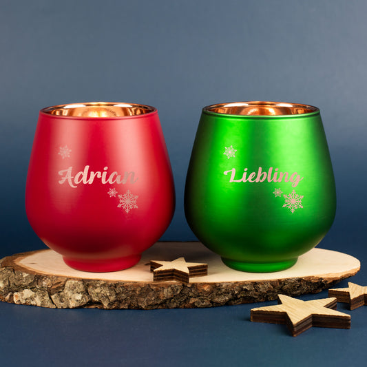 Zwei gravierte Windlichter in Rot und Grün mit Schneeflockenmotiv und Namen, auf Holzscheibe mit Sternen dekoriert.