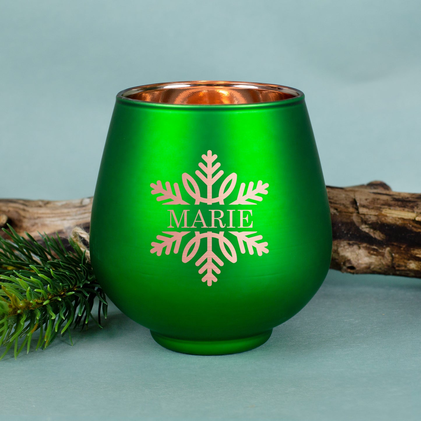 Grünes Windlicht mit Gravur „Marie“ und Schneeflockenmotiv, vor Holzstück und Tannenzweig auf blauem Hintergrund