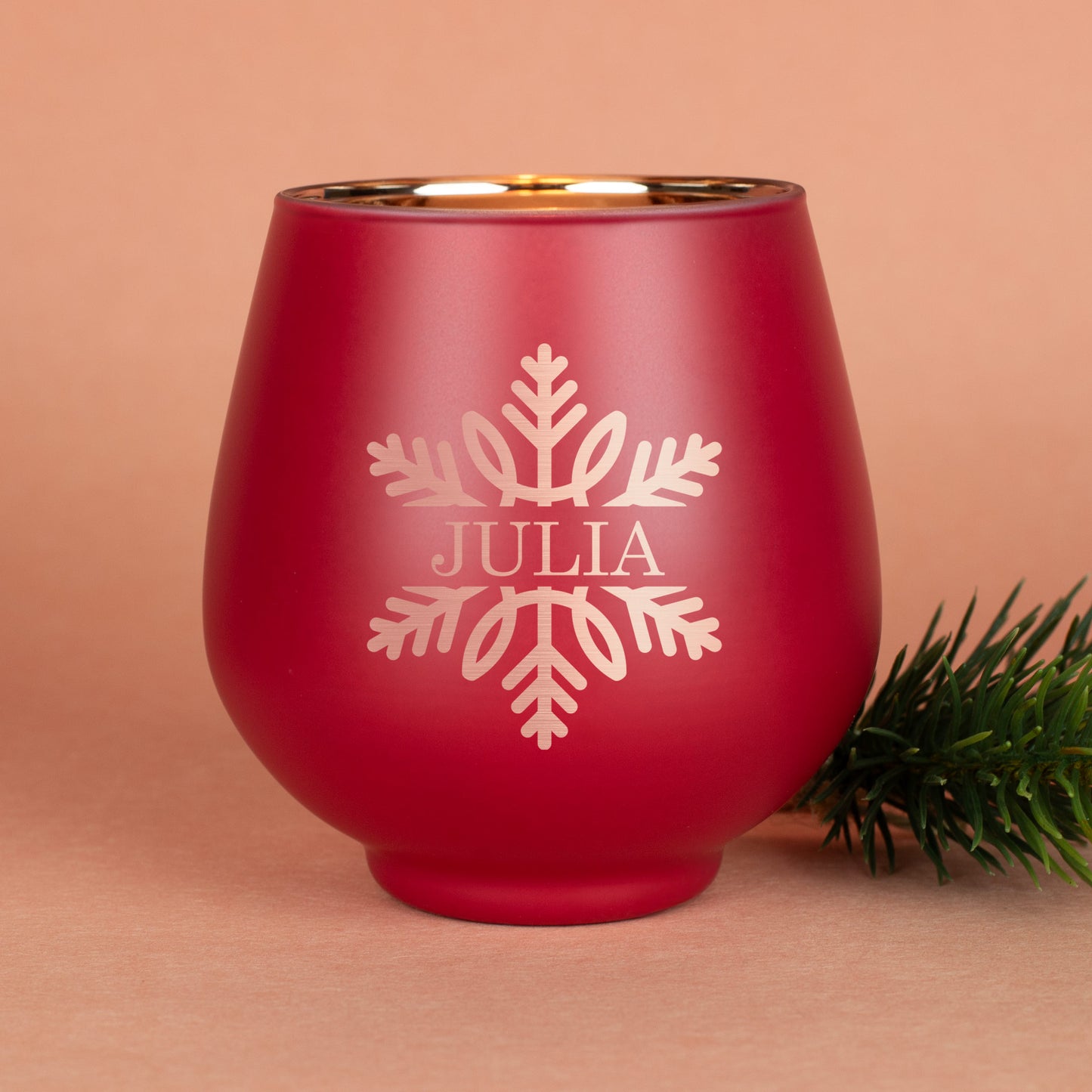 Rotes Windlicht mit Schneeflockenmotiv und Gravur „Julia“, daneben Tannenzweig auf beigem Hintergrund