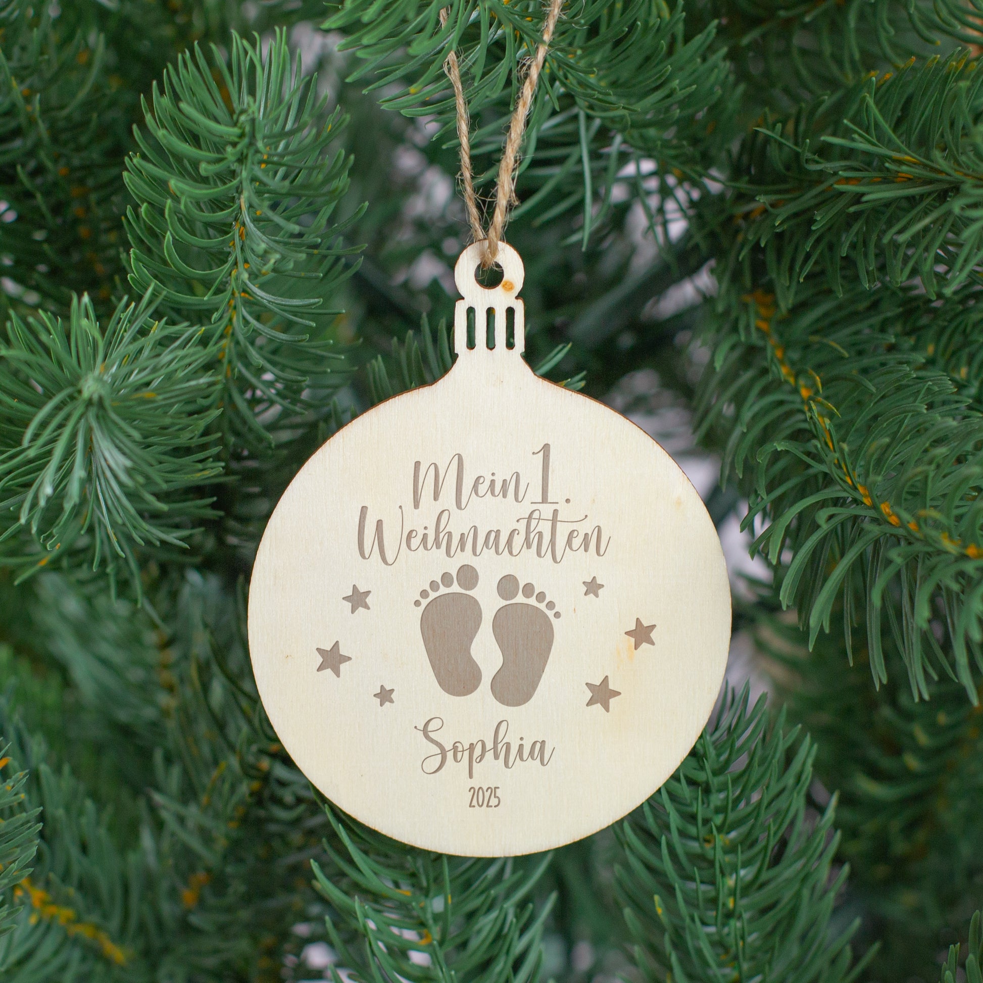 Personalisierter Holzanhänger „Mein 1. Weihnachten“ mit Gravur und Juteschnur, hängend am Weihnachtsbaum zwischen grünen Tannenzweigen.