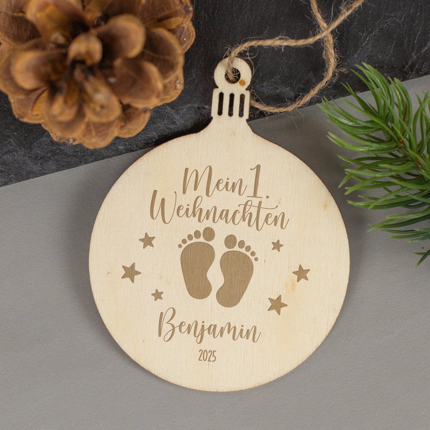 Weihnachtsanhänger aus Holz mit Babyfüßchen-Motiv und Gravur „Mein 1. Weihnachten – Benjamin 2025“, liegend neben Zapfen und Zweig.