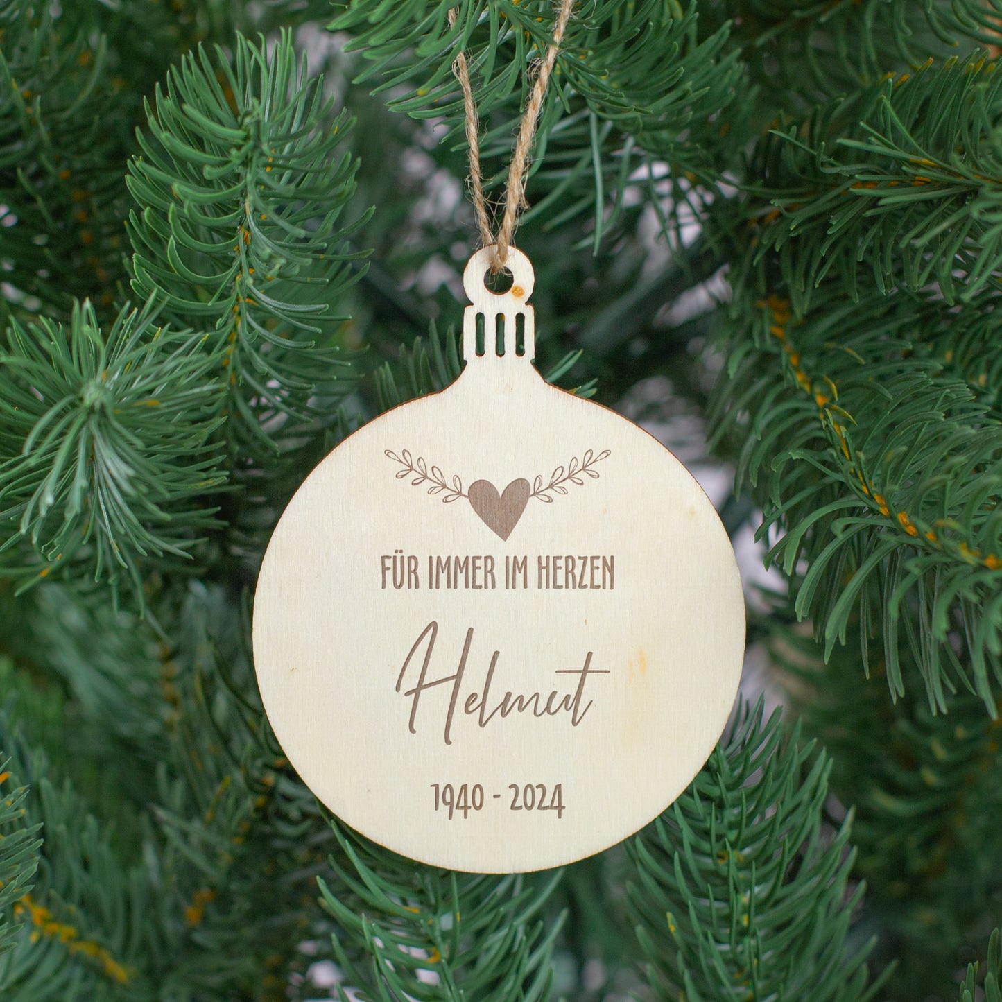 Erinnerungsanhänger aus Holz mit Gravur „Für immer im Herzen“, Name Helmut und Jahreszahlen 1940–2024, hängend am Weihnachtsbaum zwischen grünen Zweigen.