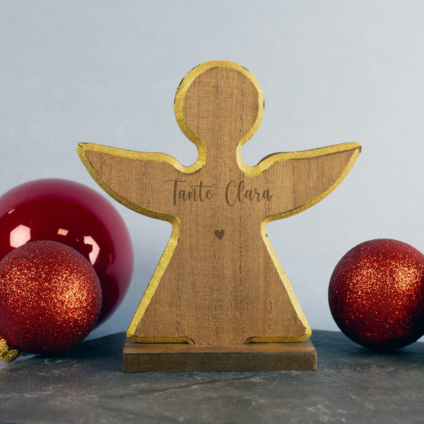 Holzengel mit ausgebreiteten Flügeln und Gravur „Tante Clara“ unter einem kleinen Herz, flankiert von zwei roten Weihnachtskugeln.