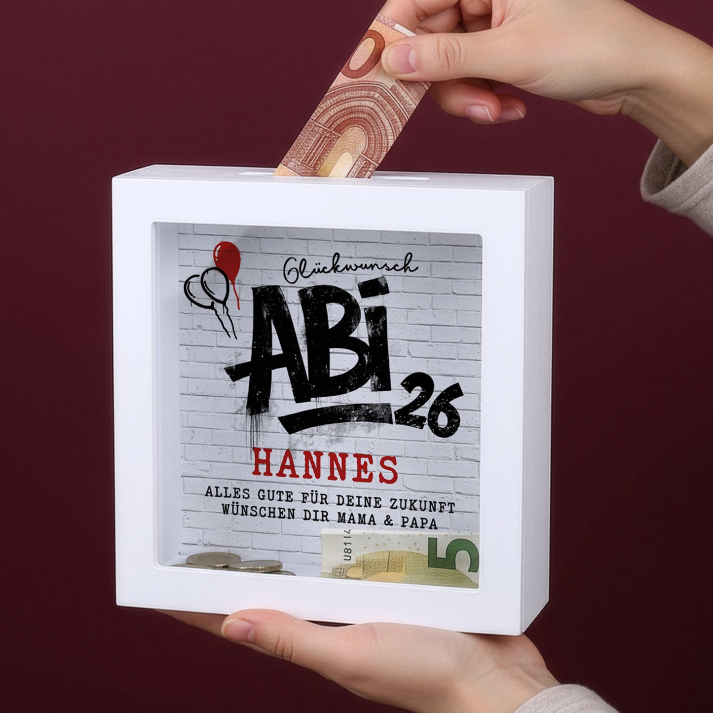 Personalisierte Spardose mit Abi 26 Motiv und Name Hannes, in den Händen gehalten und mit Geldschein befüllt
