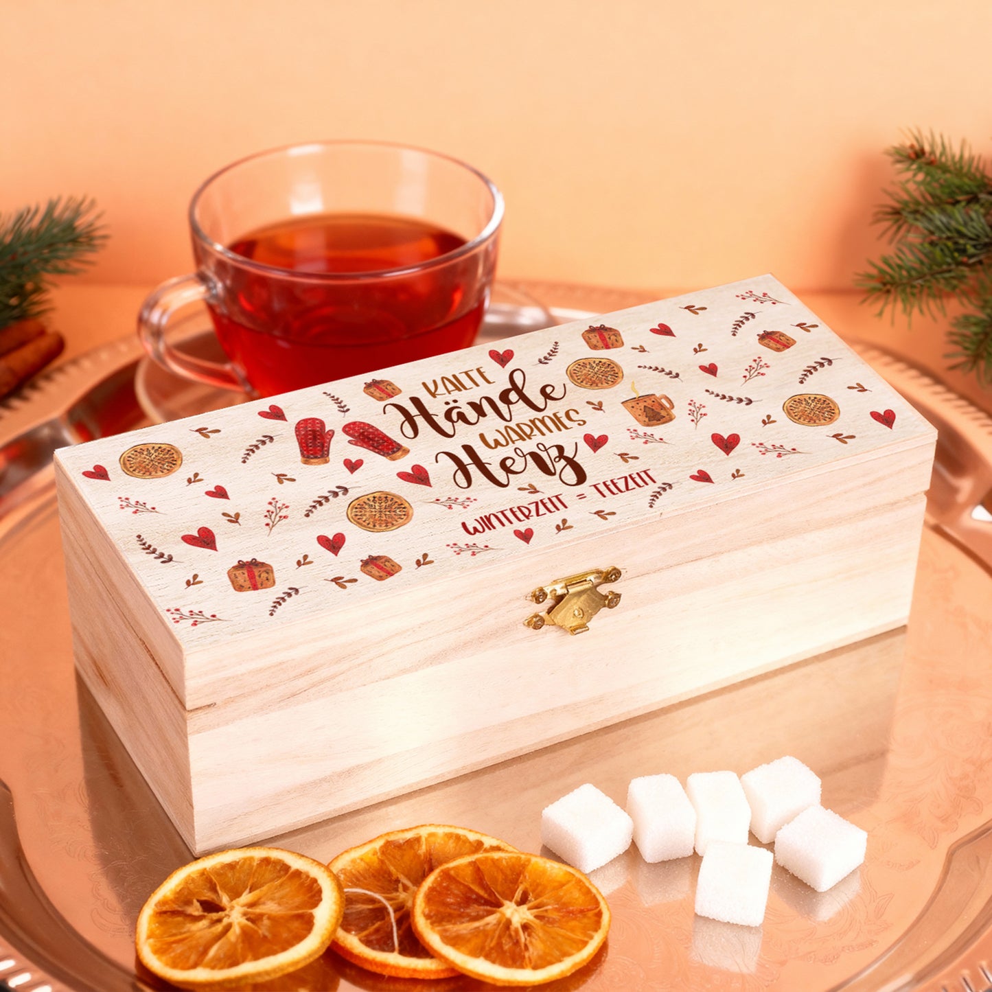 Personalisierte Teebox mit Gravur „Kalte Hände, warmes Herz – Winterzeit = Teezeit“, arrangiert auf einem Tablett mit Teetasse, Zuckerwürfeln und getrockneten Orangenscheiben, warmes Licht.