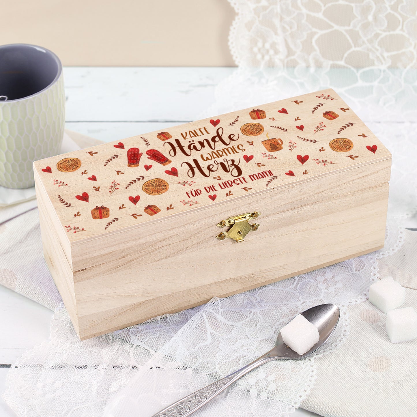 Holz-Teebox mit Gravur „Kalte Hände, warmes Herz“ und Wunschtext „Für die liebste Mami“, dekoriert mit Spitzenstoff, Teetasse und Zuckerwürfeln auf hellem Hintergrund.