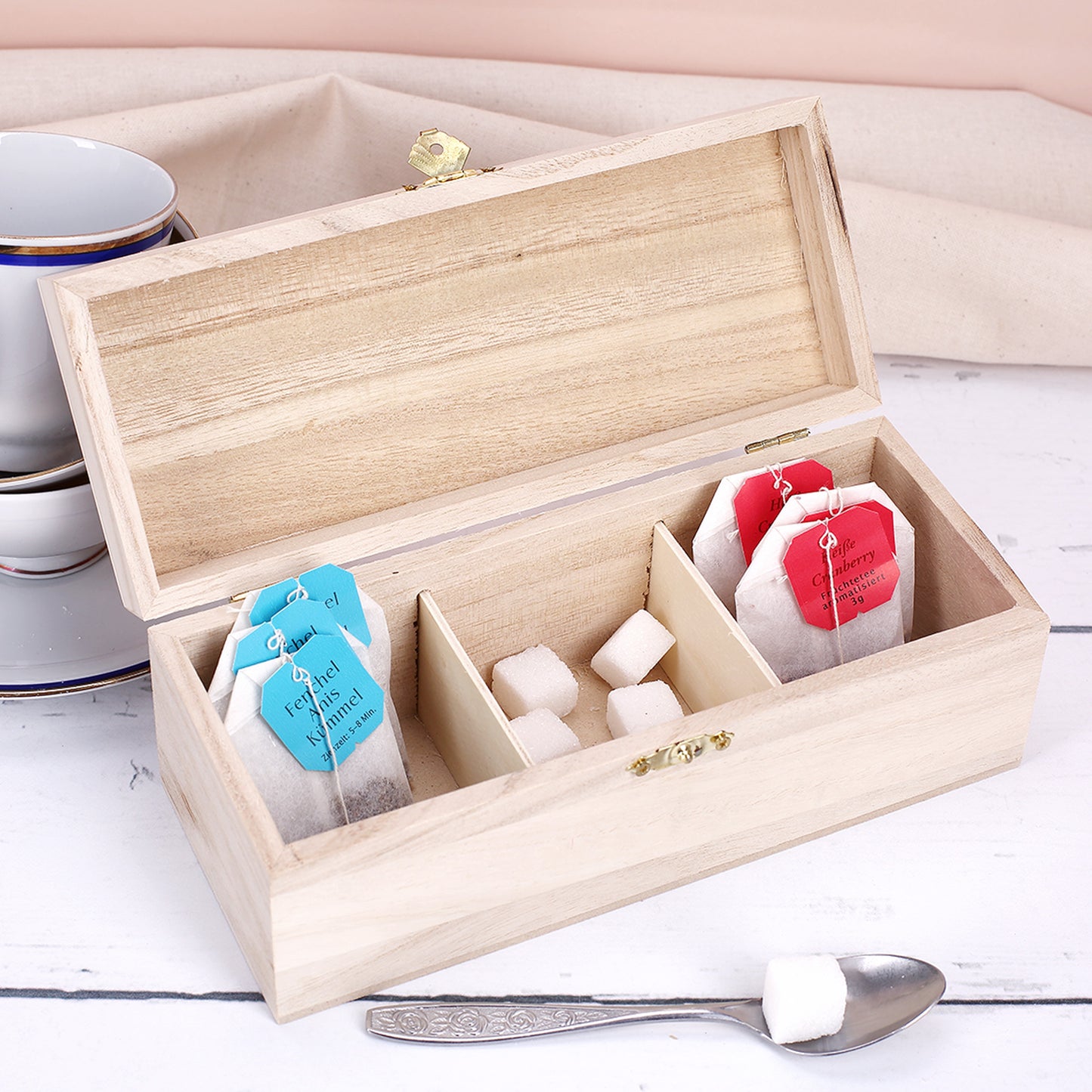 Offene Teebox aus hellem Holz mit drei Fächern, gefüllt mit Teebeuteln und Zuckerwürfeln, goldener Verschluss vorne sichtbar.