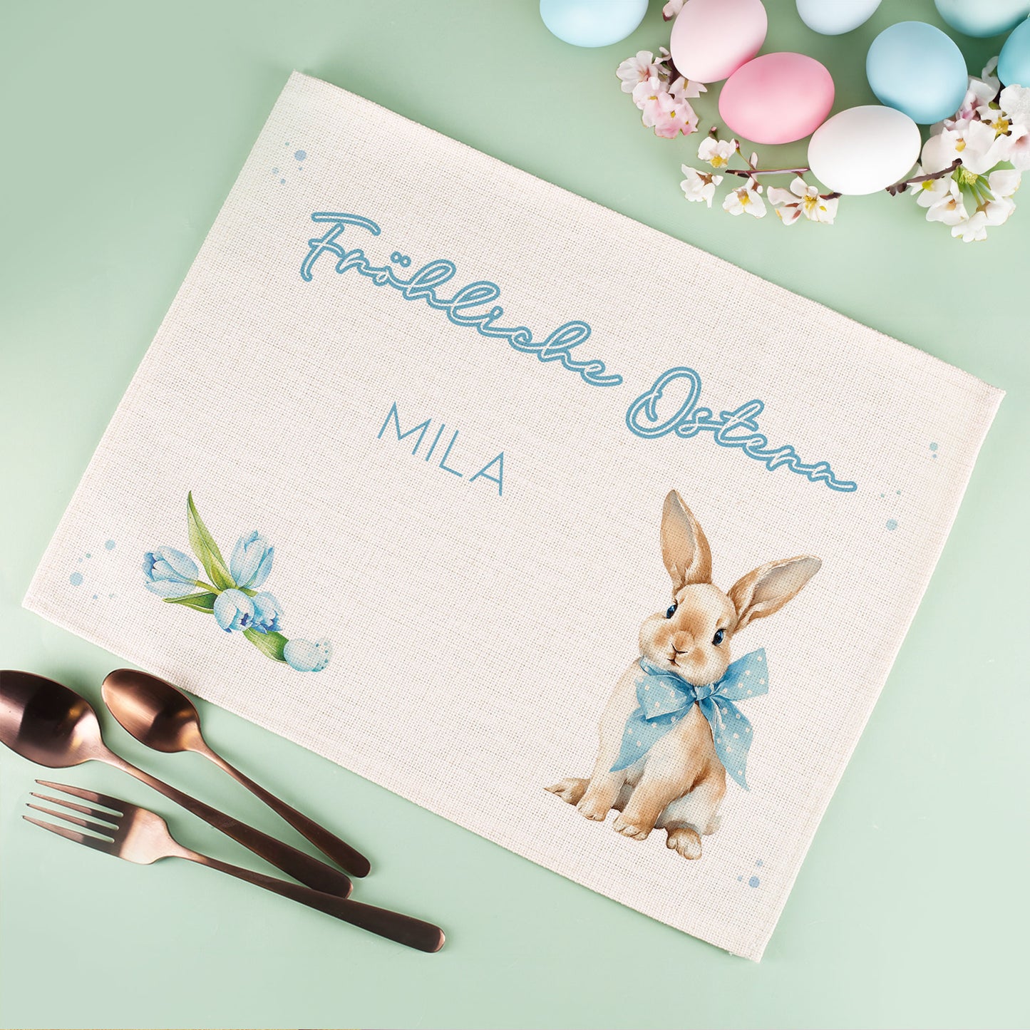 Jute Tischset zu Ostern mit Schriftzug Frohe Ostern, Osterhasenmotiv und Wunschtext auf dekoriertem Ostertisch