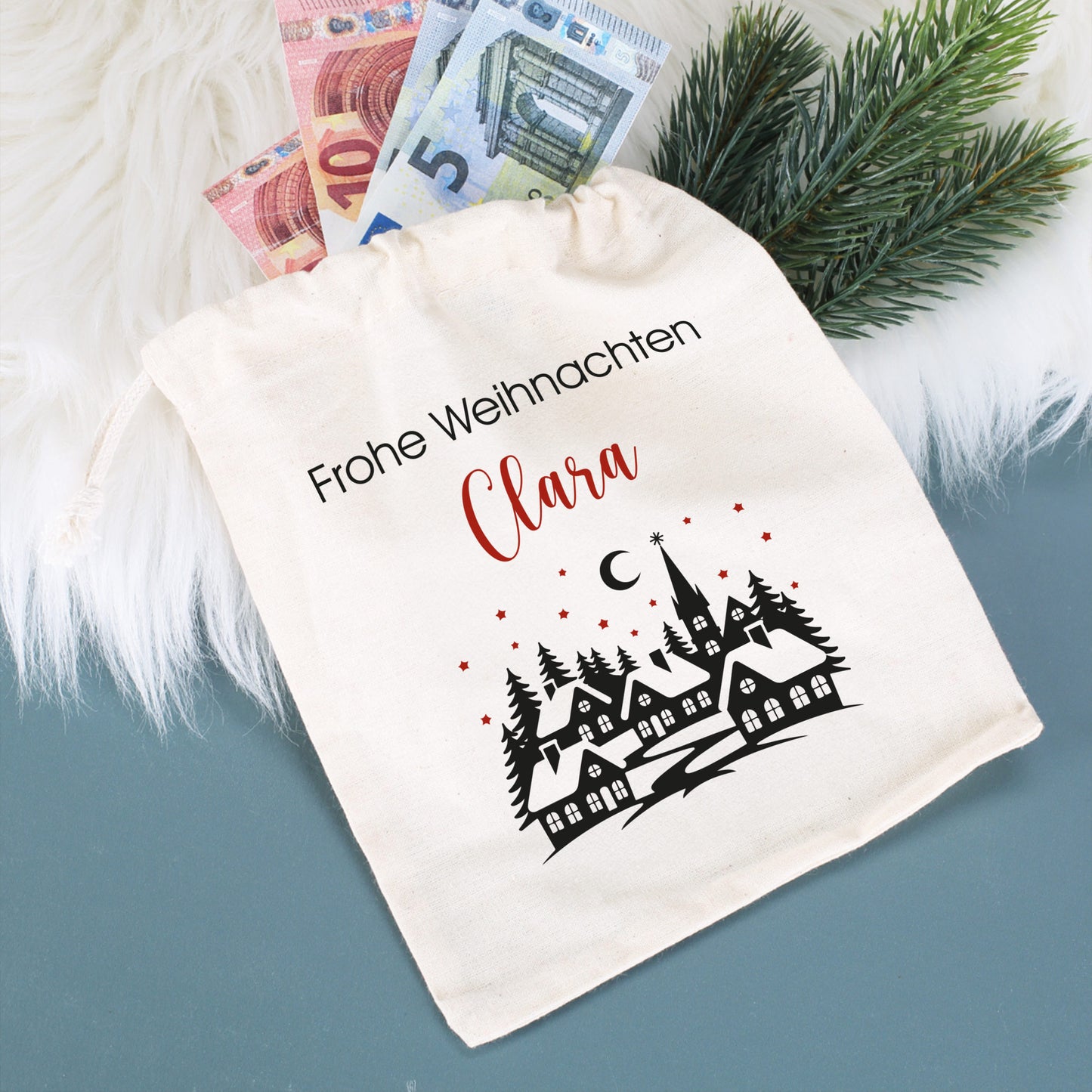 Kleiner Geschenksack mit Aufdruck „Frohe Weihnachten Clara“, befüllt mit Geldscheinen und umrahmt von Tannengrün und weißem Fell.