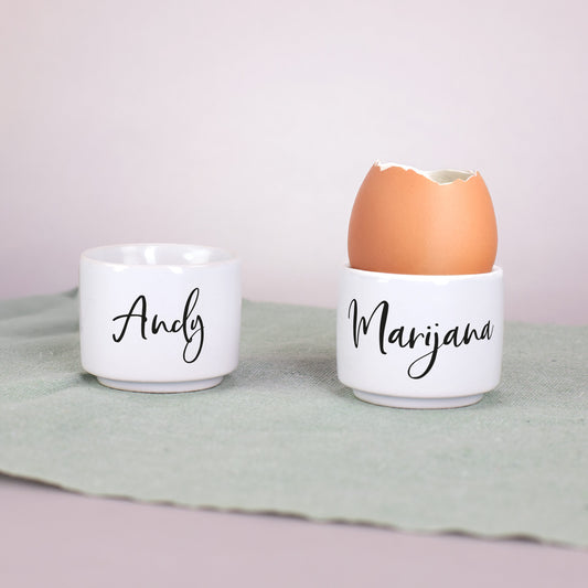 Zwei weiße Eierbecher mit den Namen „Andy“ und „Marijana“ in schwarzer Schrift, auf grünem Stoff mit Ei im Becher