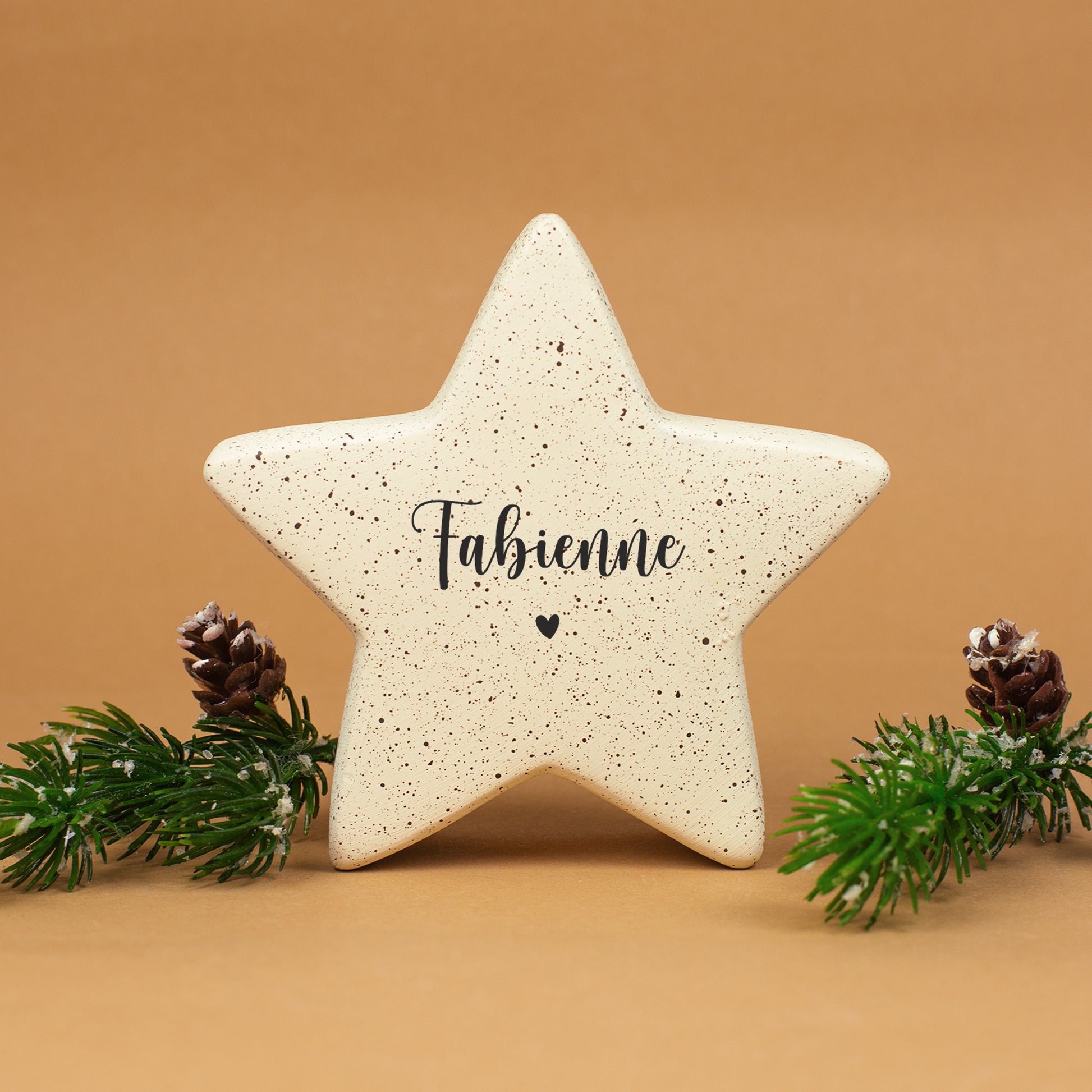 Kleiner cremefarbener Keramikstern mit dem Namen „Fabienne“ und Herzmotiv, arrangiert mit Tannenzweigen und Zapfen.