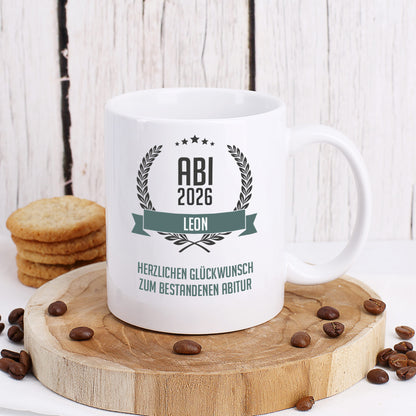 Glückwunsch-Tasse zum Abitur mit Name und Wunschtext dekoriert auf Baumscheibe mit Keksen und Kaffeebohnen