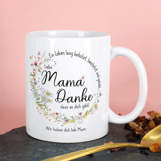 Personalisierte weiße Keramiktasse mit seidenglänzendem Finish. Das Motiv zeigt einen kreisförmigen Blumenkranz aus Wiesenblumen mit dem Spruch „Ein Leben lang behütet, beschützt und geliebt…“. In der Mitte steht „Liebe Mama, danke, dass es dich gibt!“. Darunter ein individueller Wunschtext. Perfektes Geschenk zum Muttertag, Geburtstag oder als liebevolle Aufmerksamkeit.