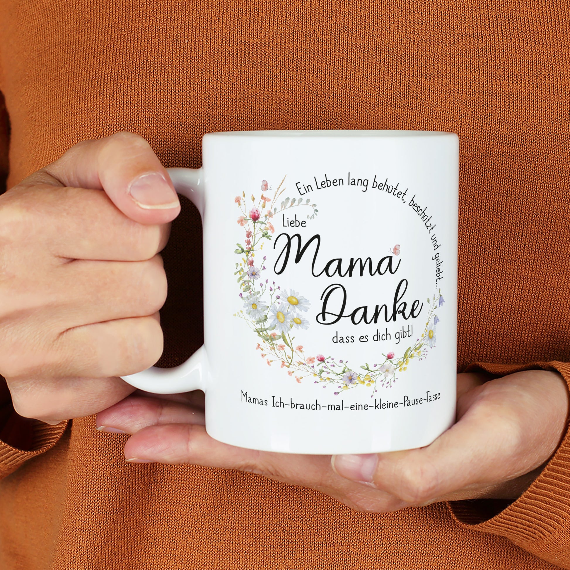 Weiße Keramiktasse mit seidenglänzendem Finish, bedruckt mit einem Wiesenblumen-Kranz und dem Spruch „Ein Leben lang behütet, beschützt und geliebt…“. In der Mitte steht „Liebe Mama, danke, dass es dich gibt!“, ergänzt durch einen individuellen Wunschtext. Die Tasse wird von einer Person in einem warmen, orangefarbenen Pullover gehalten. Ein liebevolles Geschenk für Mütter zum Muttertag, Geburtstag oder als Dankeschön.