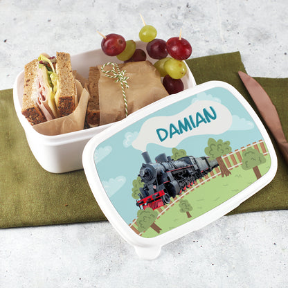 Personalisierte Brotdose mit Retro-Dampflok-Motiv und dem Namen „Damian“, geöffnet mit belegten Broten und Trauben, auf grünem Tuch arrangiert.