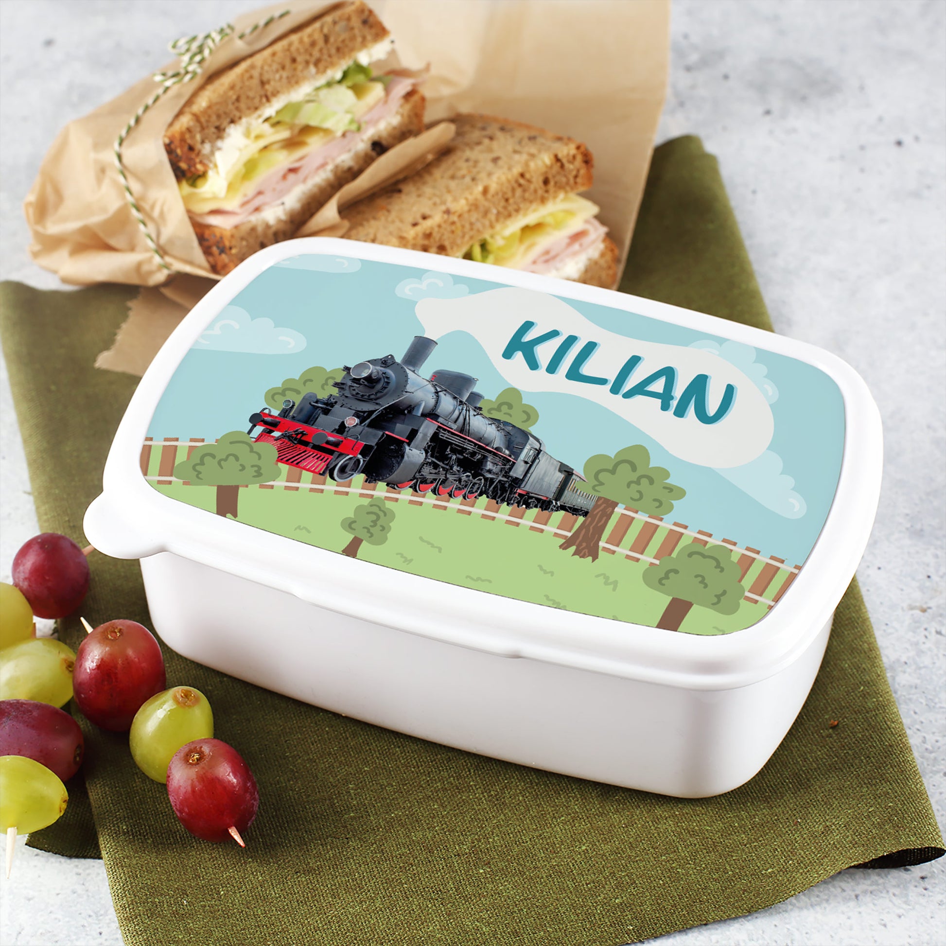 Brotdose mit Eisenbahnmotiv und dem Namen „Kilian“ auf dem Deckel, daneben Sandwiches und Trauben als Schulpausen-Snack.