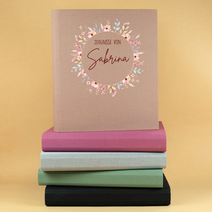 Personalisierte Zeugnismappe aus Leinen in Beige mit Blumenkranz und Name Sabrina auf einem Stapel farbiger Dokumentenmappen.