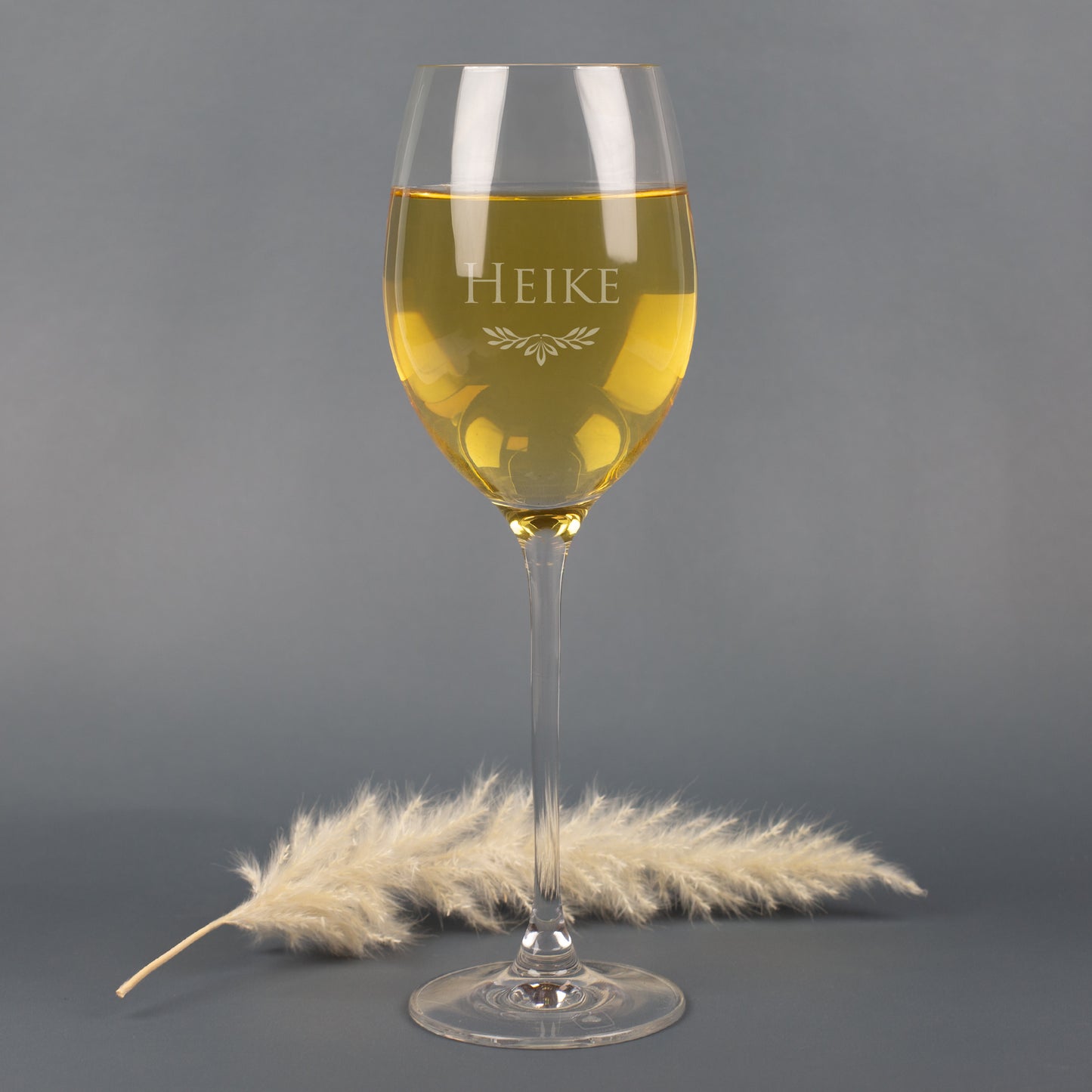 Leonardo Weißweinglas mit Gravur Name Heike und Ornamentmotiv, gefüllt mit Weißwein, auf grauem Hintergrund mit Pampasgras