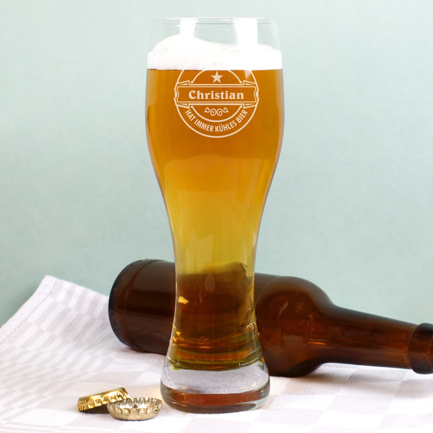 Weizenglas mit Gravur „Christian hat immer kühles Bier“, gefüllt mit Bier, daneben liegende braune Flasche und Kronkorken auf weißem Tuch.