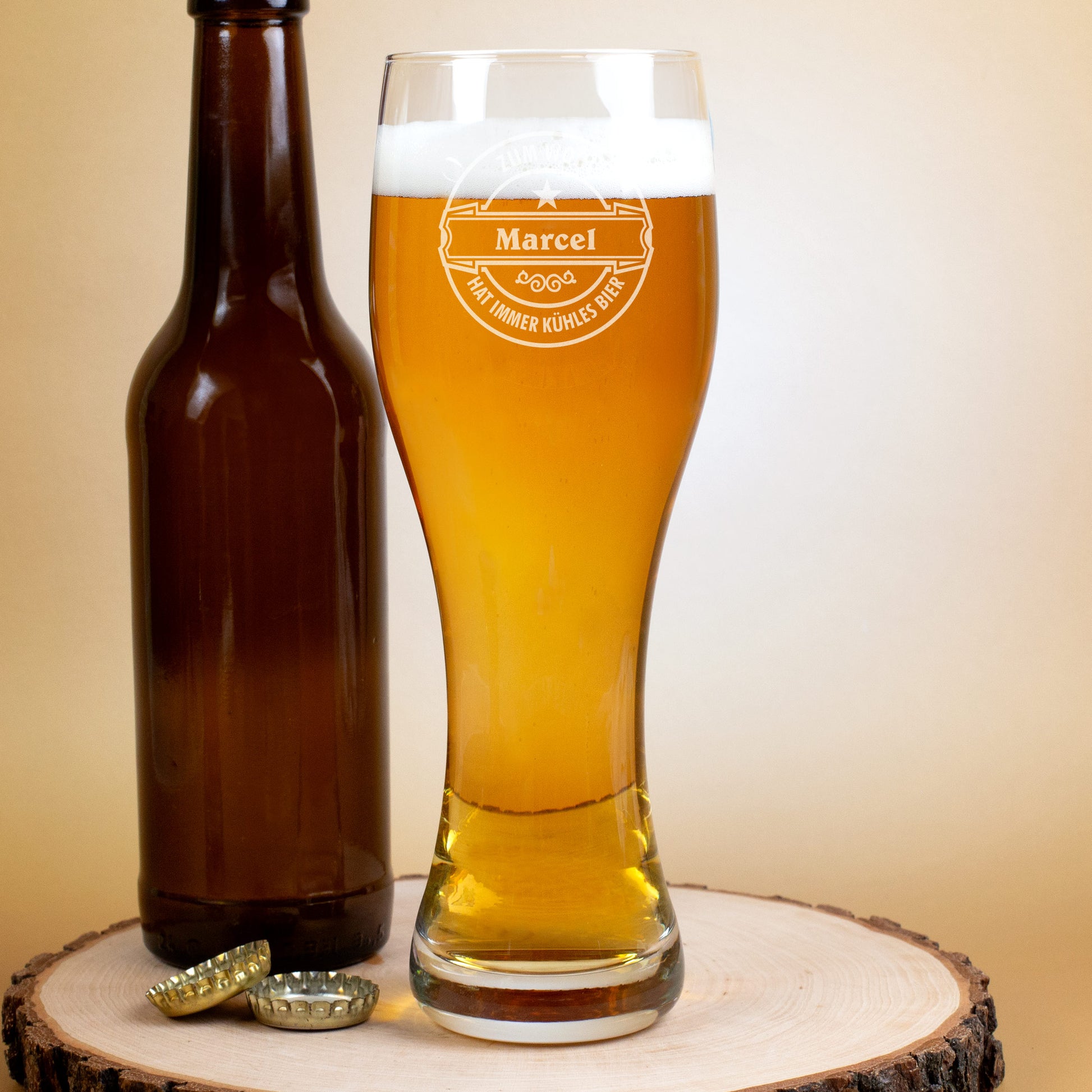 Weizenglas mit Gravur „Marcel hat immer kühles Bier“, gefüllt mit Bier, daneben braune Flasche und zwei Kronkorken auf Holzscheibe.