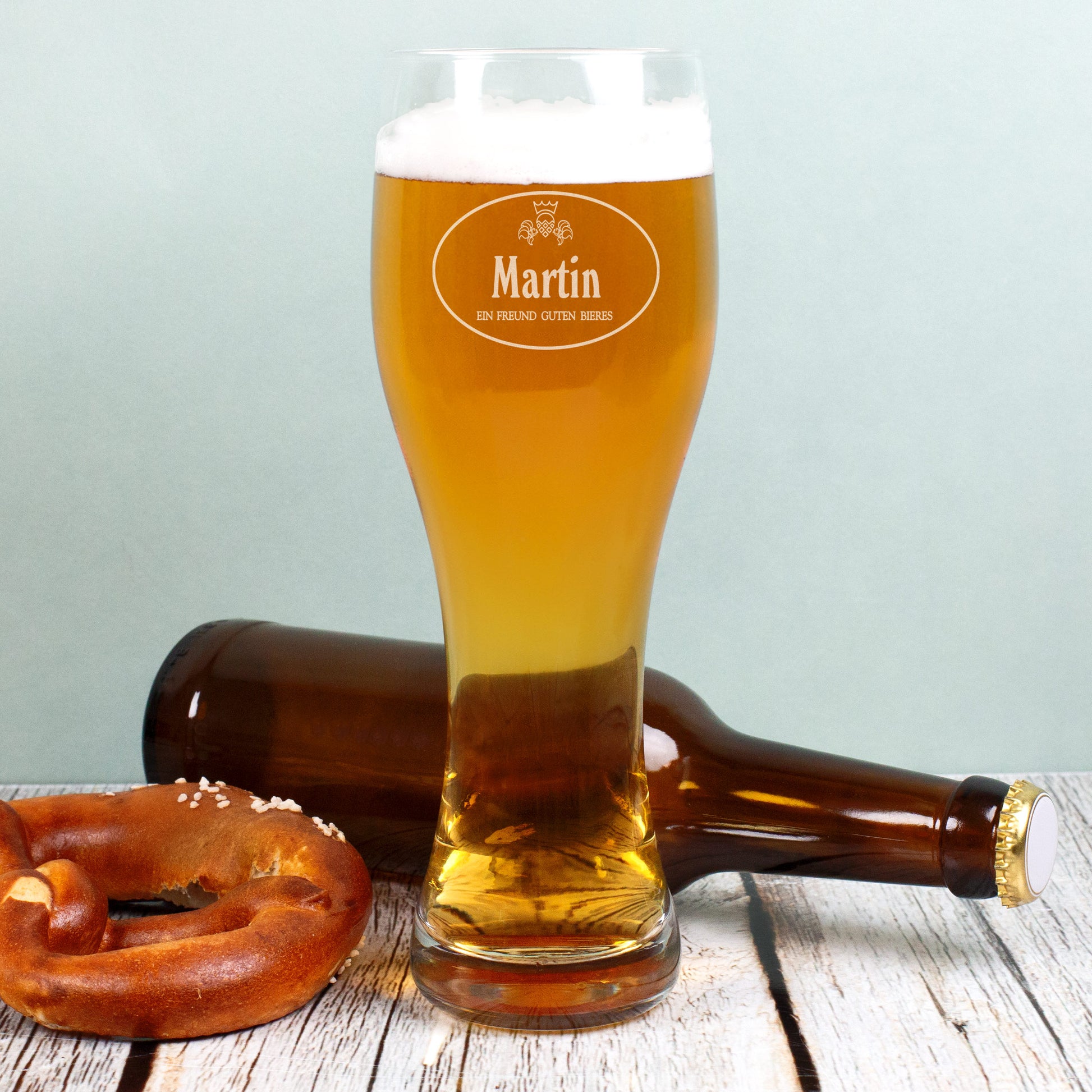 Gefülltes Weizenglas mit Gravur Martin, daneben Bierflasche und Brezel