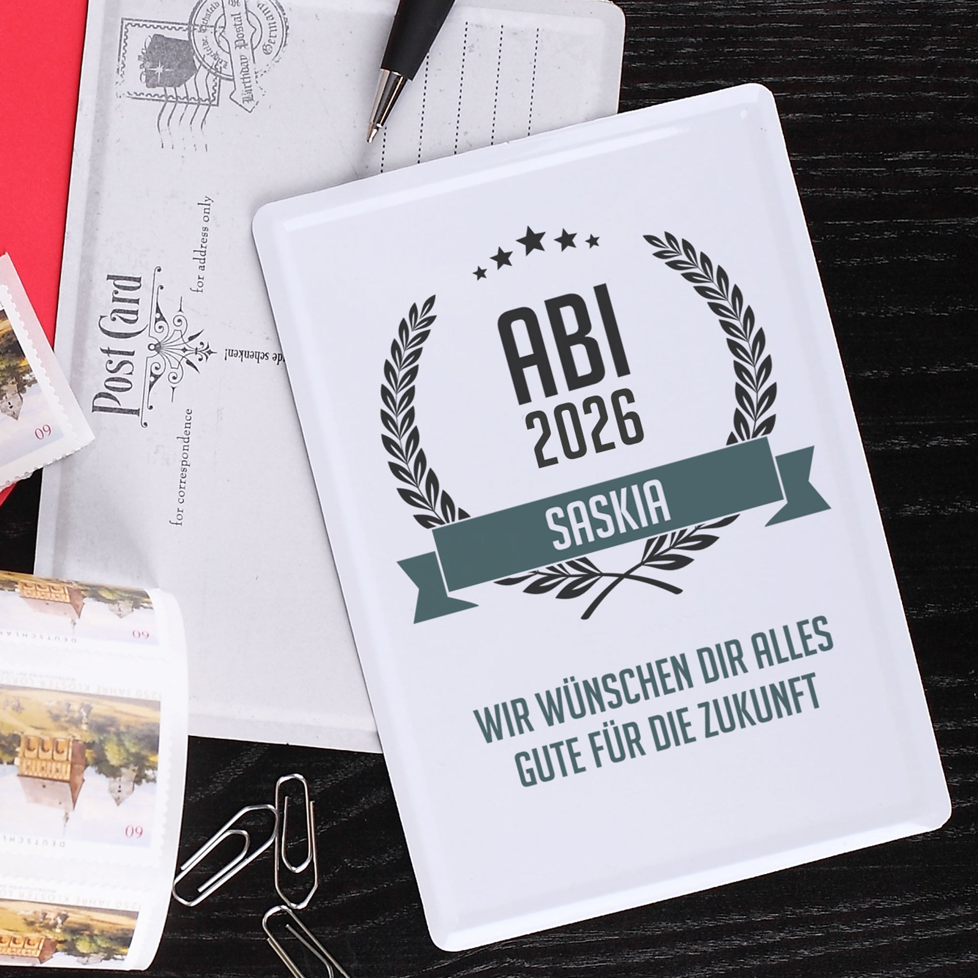 Abitur Blechpostkarte mit Glückwunschtext und Name auf dunklem Hintergrund mit Briefmarken und Büroklammern