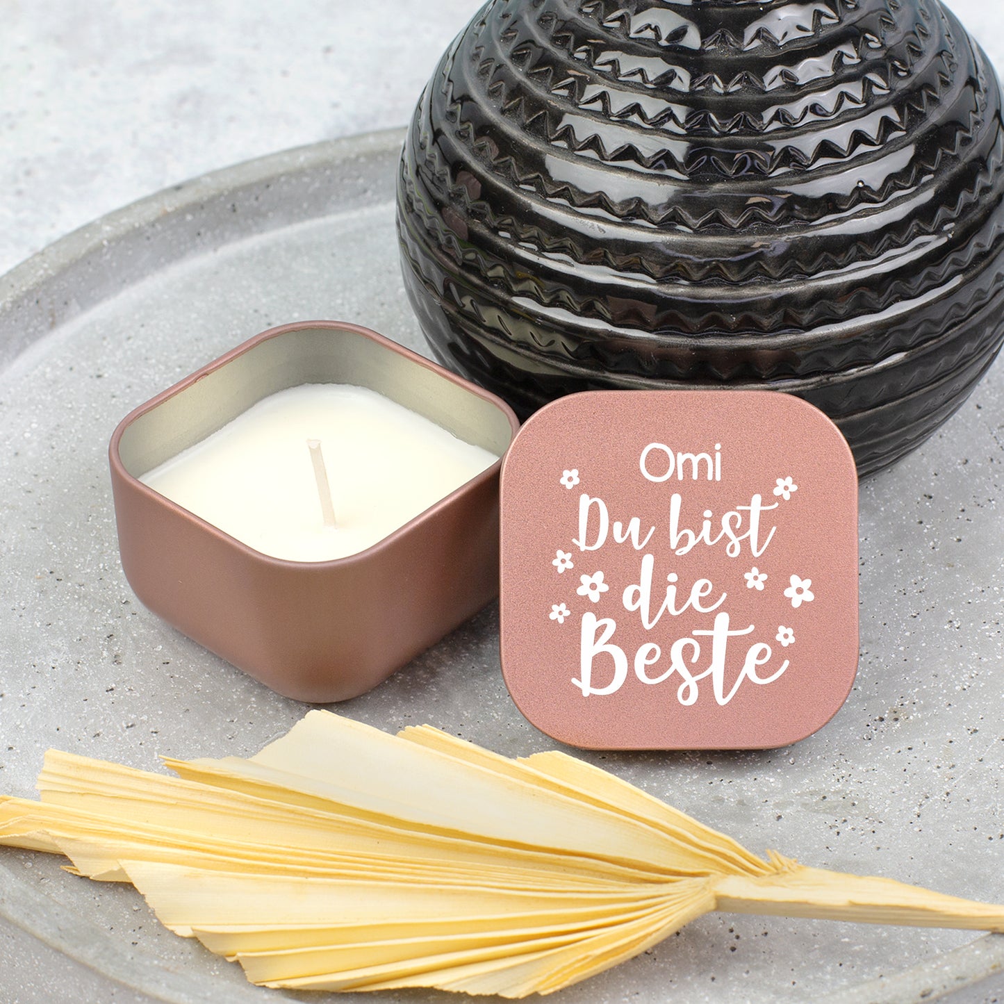 Roségoldene Duftkerze mit Vanilleduft neben schwarzer Vase, Deckel mit Aufschrift „Omi Du bist die Beste“