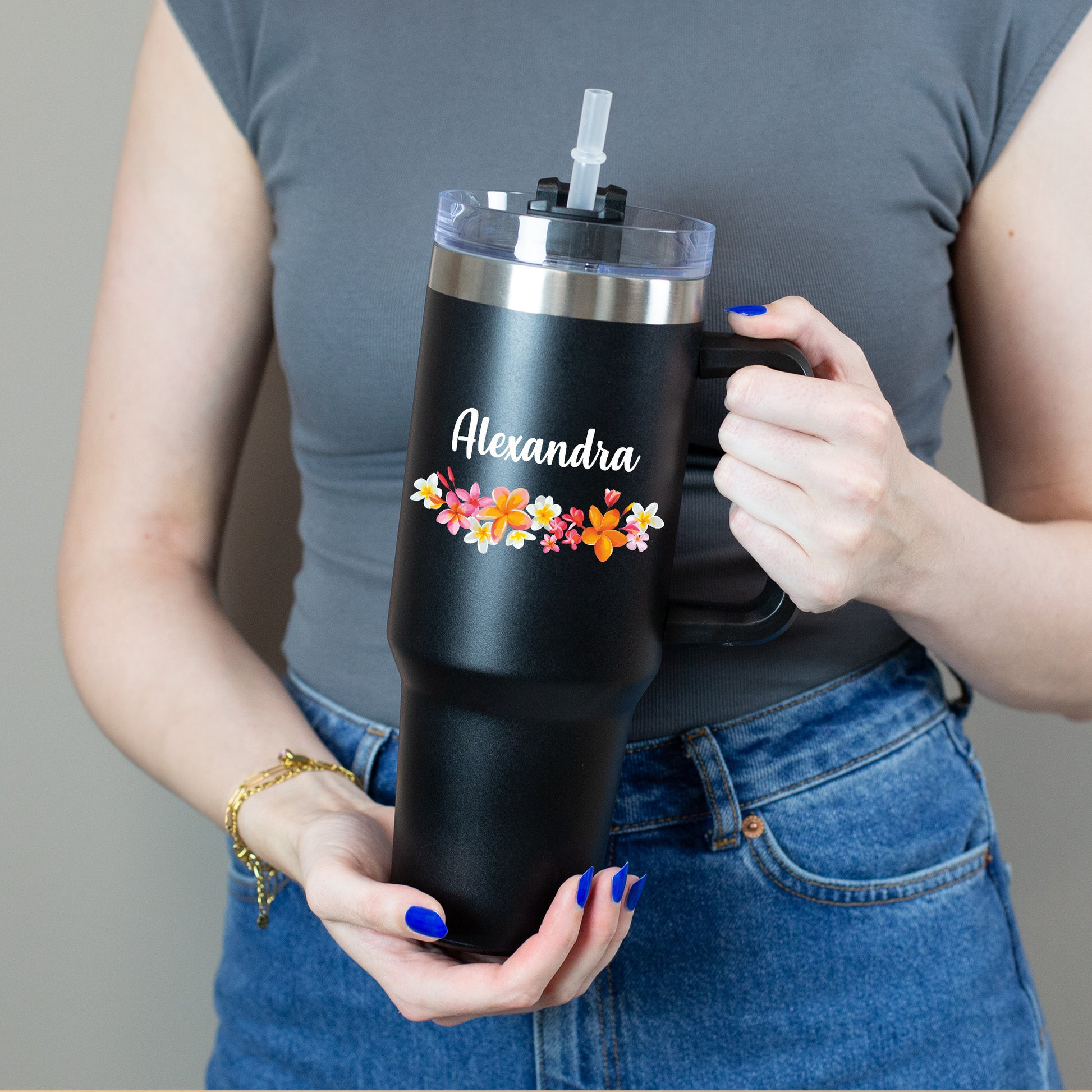 Person hält  Thermobecher mit buntem Blumenkranz und weißem Namen mit beiden Händen