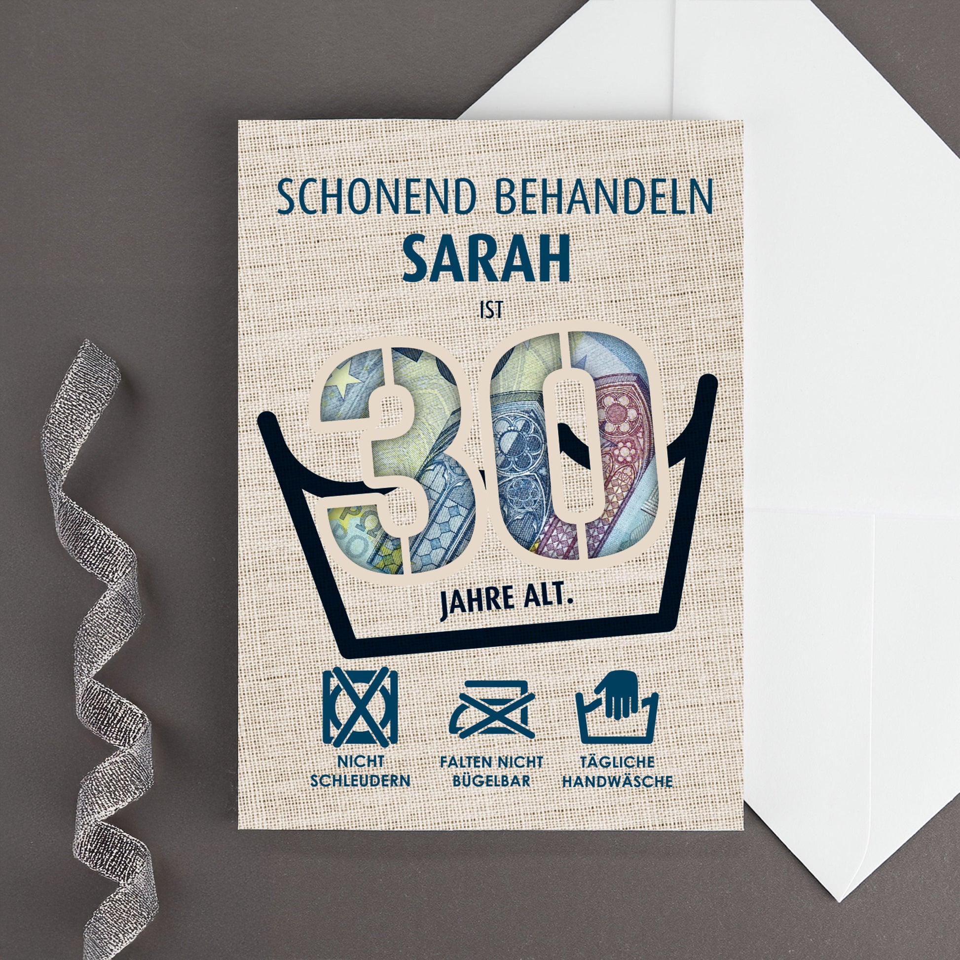 Klappkarte zum 30. Geburtstag mit dem Namen Sarah und ausgestanzter 30, hinterlegt mit Euro-Scheinen, Wäschezeichen und weißem Umschlag auf grauem Hintergrund