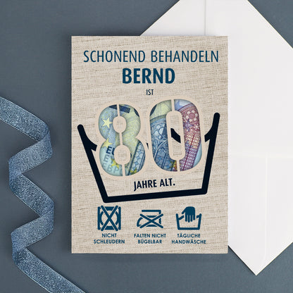 Personalisierte Geburtstagskarte mit Name „Bernd“ und sichtbarem Geldschein durch die Zahl 80, daneben ein weißer Umschlag auf dunkelblauem Hintergrund