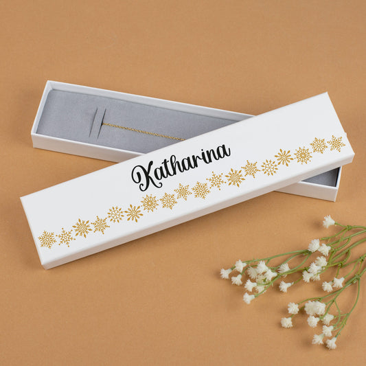 Weiße Schmuckbox mit goldenen Schneeflocken und Personalisierung „Katharina“. Die geöffnete Box zeigt den grauen Schaumstoff für Armbänder. Präsentiert vor warmem Hintergrund mit zarten weißen Blumen als Dekoration.
