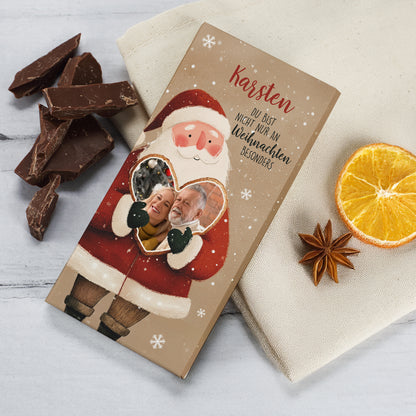 Personalisierte Weihnachts-Schokolade mit Weihnachtsmann und Herzmotiv. Auf der Verpackung ist der Name „Karsten“ aufgedruckt, darunter der Spruch „Du bist nicht nur an Weihnachten besonders“. Im Herzfenster befindet sich ein persönliches Foto. Neben der Schokolade liegen Schokoladenstücke, getrocknete Orangenscheiben und Sternanis – festlich dekoriert auf hellem Hintergrund.