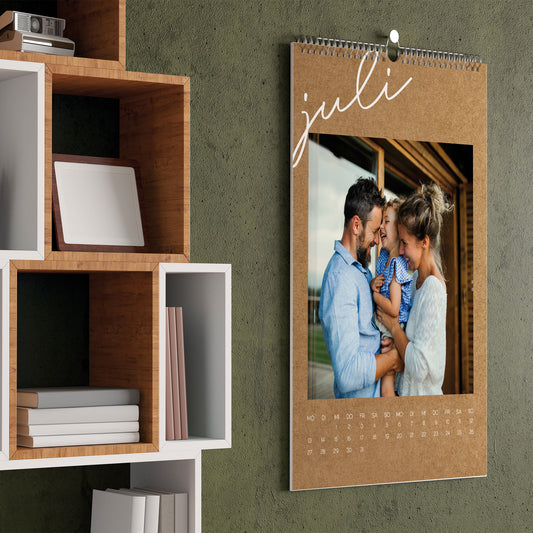 Foto-Wandkalender in Kraftpapier-Optik für den Monat Juli, mit Familienfoto und feiner Spiralbindung, aufgehängt an einer grünen Wand.