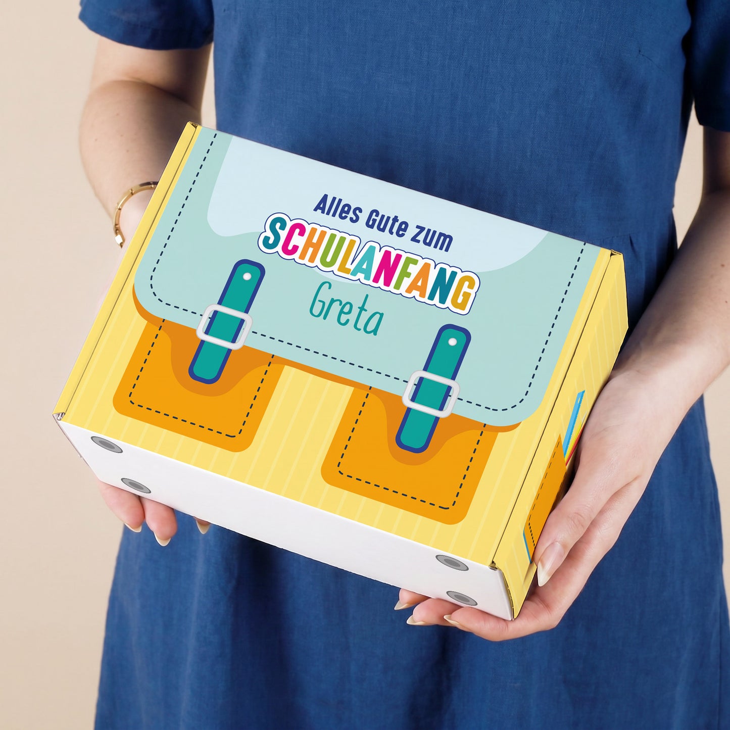 Kleine Aufbewahrungsbox aus Pappe zur Einschulung in Gelb mit Schulranzen-Design und Wunschname, in den Händen einer Person