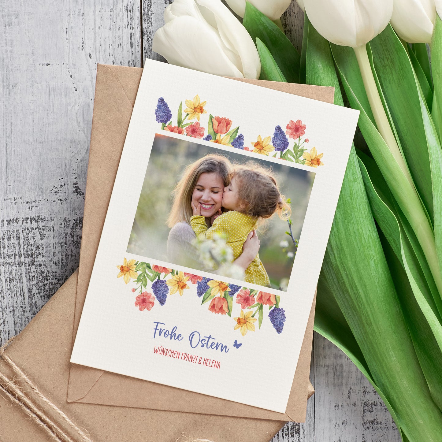 Osterkarte mit Foto und Blumenmotiv neben Umschlag und Tulpen dekorativ arrangiert
