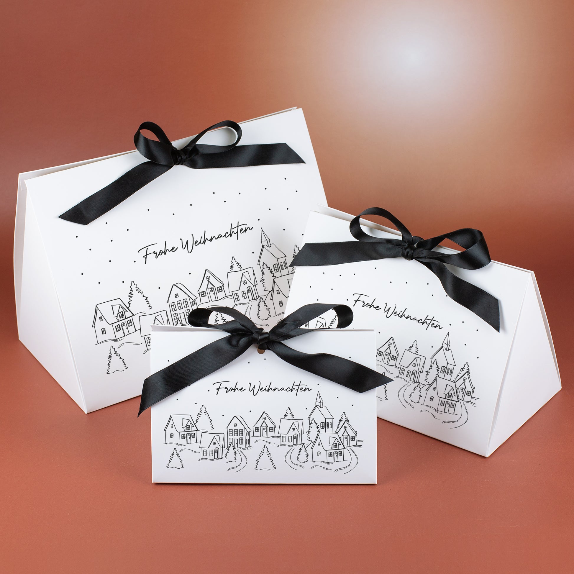 Drei Geschenktaschen in klein, mittel und groß mit Winterdorf-Motiv, Schneepunkten und schwarzer Satinschleife, vor warmem Hintergrund arrangiert.