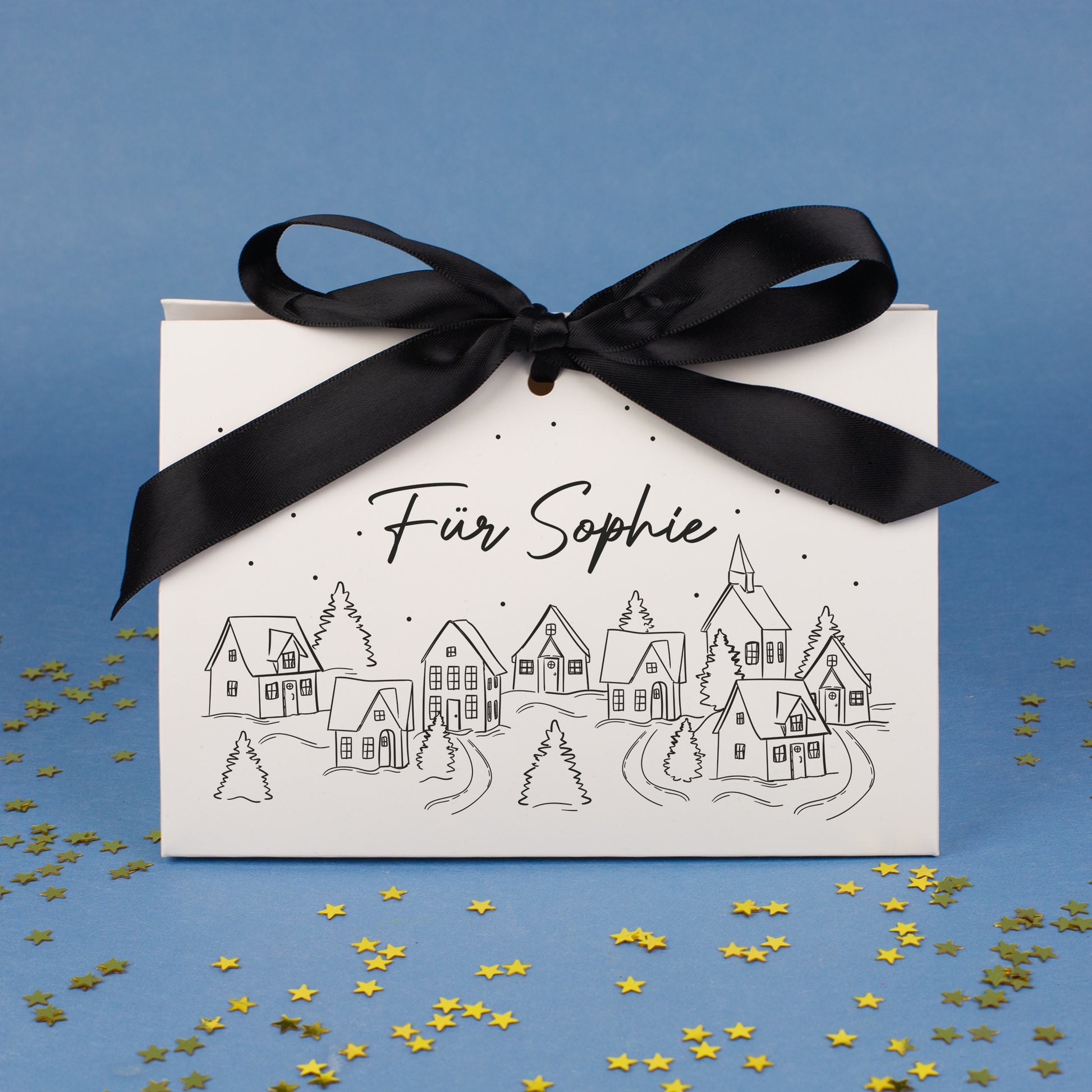 Mittlere Geschenktasche mit Winterdorf-Motiv und personalisiertem Text „Für Sophie“, dekoriert vor blauem Hintergrund mit goldenen Sternkonfetti.