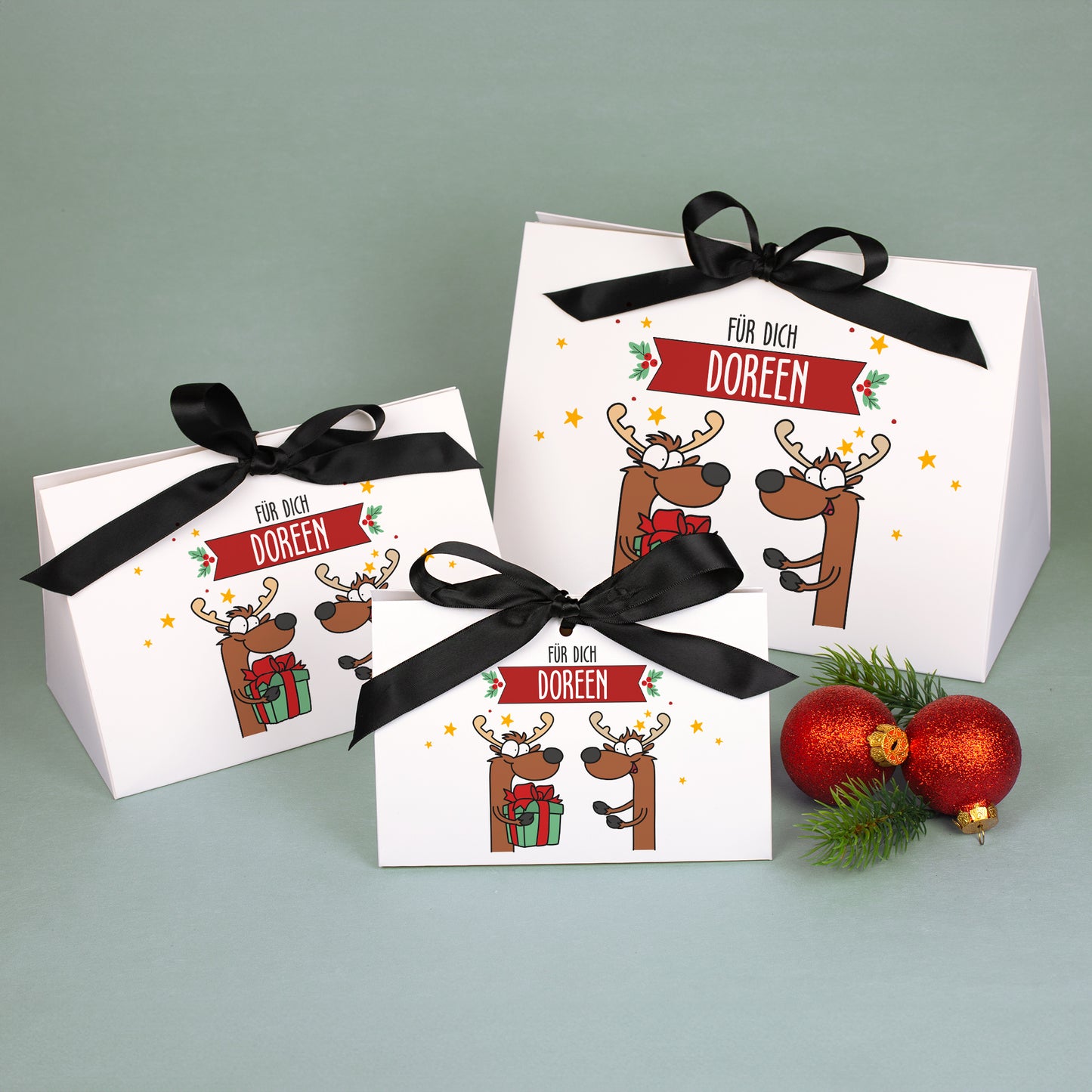 Drei Geschenktaschen in klein, mittel und groß mit Rentiermotiv und Namensbanner „Doreen“, aus weißem Karton mit schwarzer Satinschleife, dekoriert mit Weihnachtskugeln.