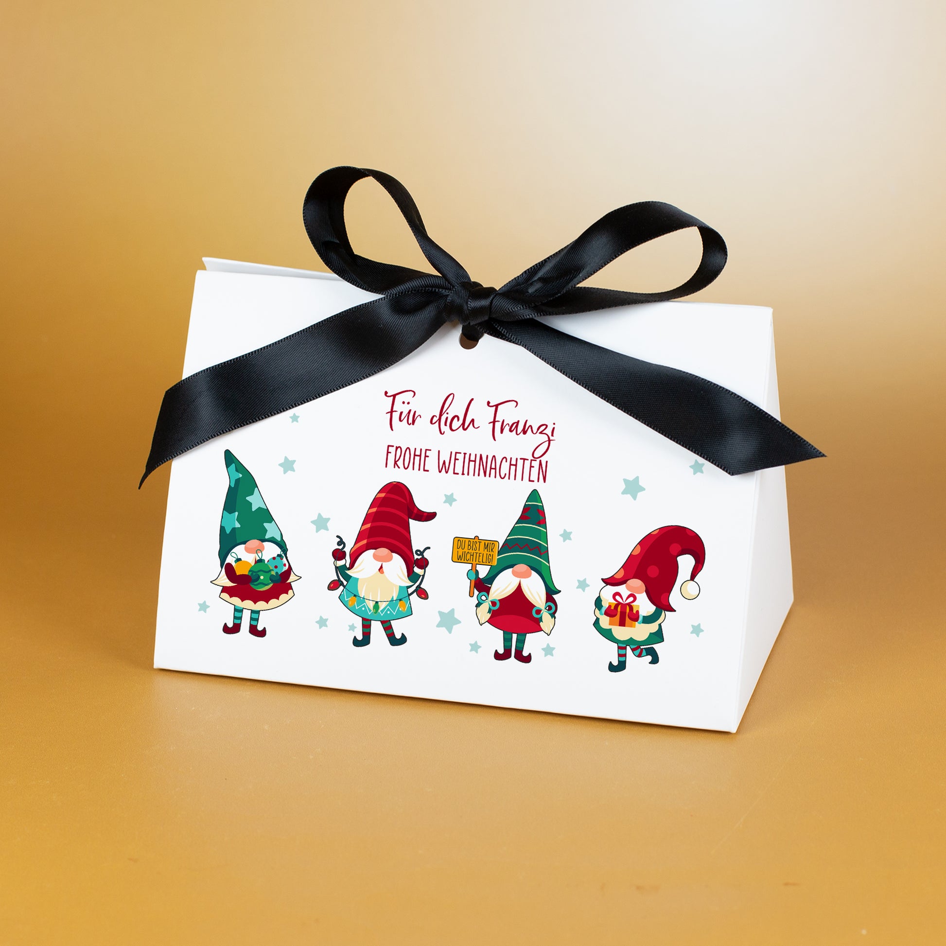 Weihnachtliche Geschenkverpackung in mittlerer Größe aus weißem Karton mit groß gebundener schwarzer Satinschleife. Auf der Vorderseite befinden sich vier farbenfrohe Wichtel und darüber der personalisierte Text „Für dich Franzi – Frohe Weihnachten“. Die Tasche steht vor goldfarbenem Hintergrund und zeigt die Motive klar und detailreich.