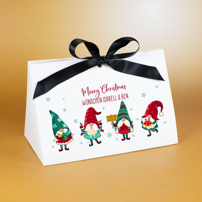 Große Geschenkverpackung zu Weihnachten mit schwarzem Satinband und farbigem Wichtelmotiv. Über dem Motiv steht der personalisierte Text „Merry Christmas wünschen Isabell & Ben“ in roter Schrift. Die vier Wichtel tragen weihnachtliche Outfits und stehen vor einem Muster kleiner Sterne auf weißem Karton. Präsentiert vor goldenem Hintergrund.