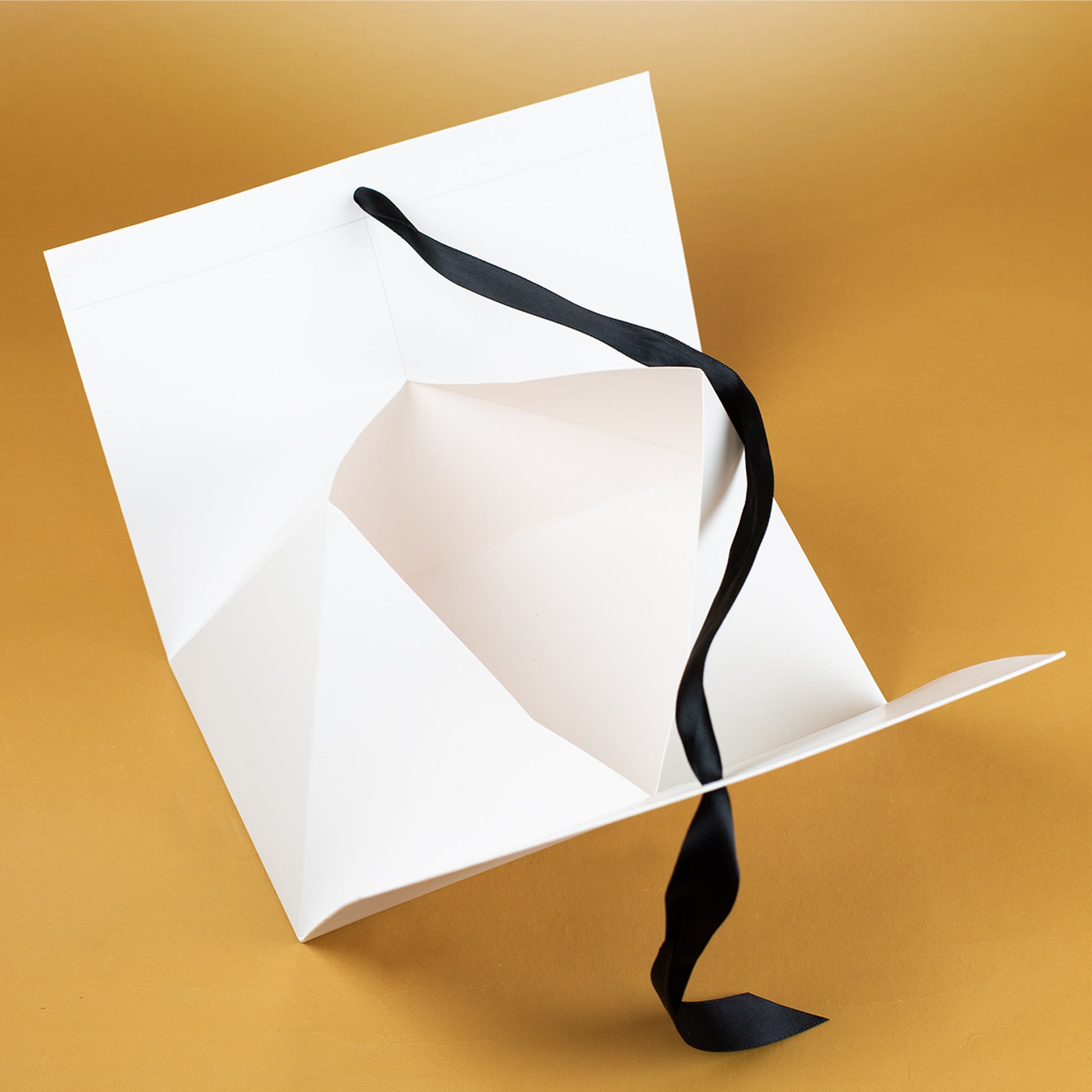 Innenansicht der Geschenkverpackung aus weißem Karton, geöffnet und flach auf goldenem Untergrund liegend. Gut erkennbar sind die stabile Faltkonstruktion, der großzügige Innenraum sowie das lose schwarze Satinband, das später zur Schleife gebunden wird. Die Aufnahme zeigt die Materialstärke und Aufbauweise der Geschenkbox.