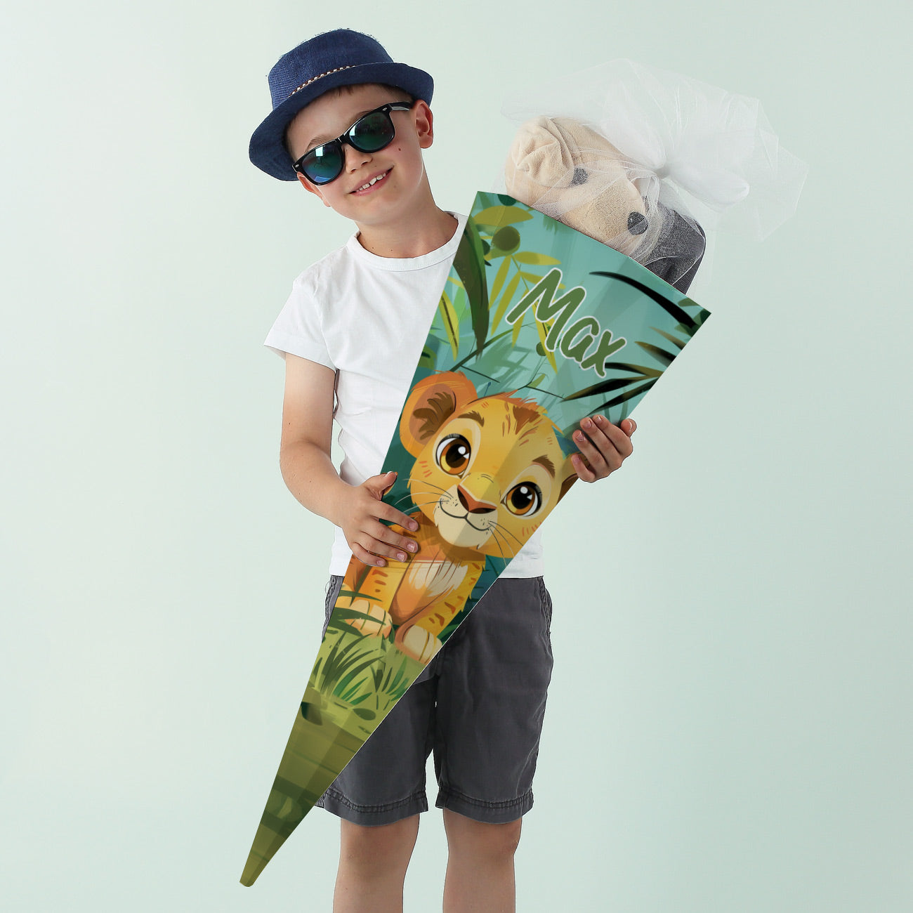 Junge mit Sonnenbrille und Hut lächelt mit einer bunten Schultüte mit Löwenmotiv und Name „Max“.