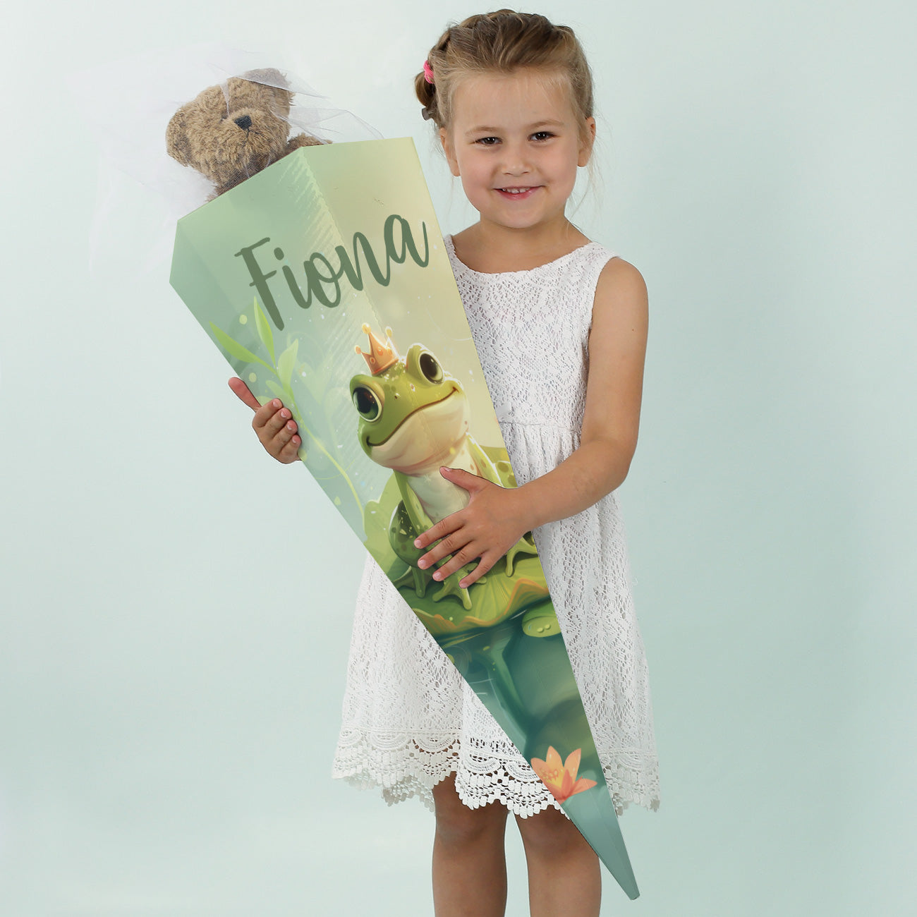 Mädchen in weißem Kleid lächelt mit einer großen Zuckertüte mit dem Namen "Fiona" und Frosch-Motiv