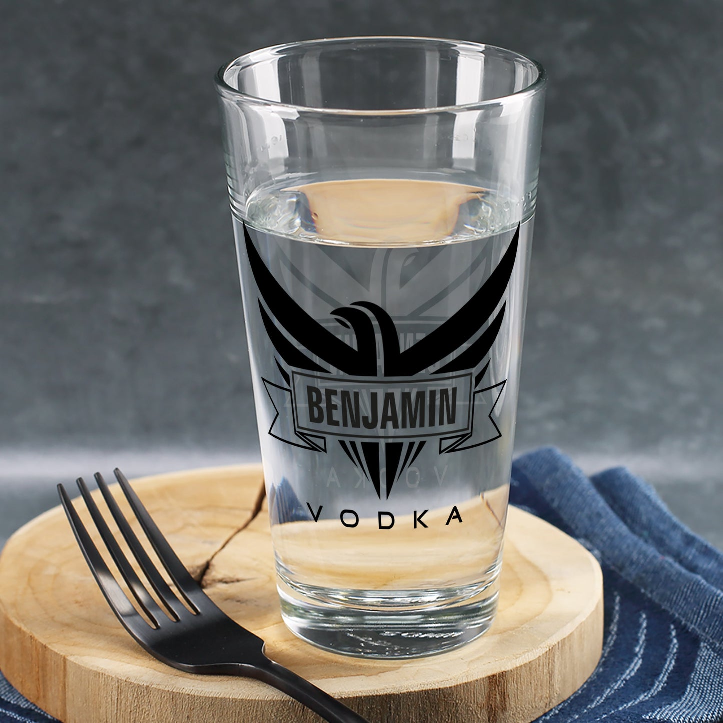 Trinkglas mit schwarzem Adler-Motiv und Schriftzug „Vodka“, personalisiert mit Name Benjamin, gefüllt mit Wasser auf Holzbrett