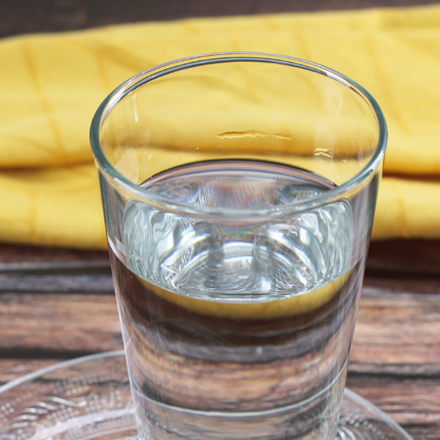 Klares Trinkglas ohne Motiv, gefüllt mit Wasser, auf Holztisch mit gelbem Stoff im Hintergrund.