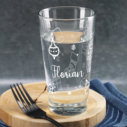 Personalisierbares Trinkglas mit Ornamenten – Stern, Strumpf und Tannenbaum – mit Wasser gefüllt und Namen, auf Holzuntergrund.