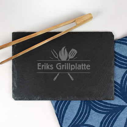 Schwarze Schieferplatte mit Gravur „Eriks Grillplatte“ und Grillutensilien, Holz-Grillzange als Dekoration