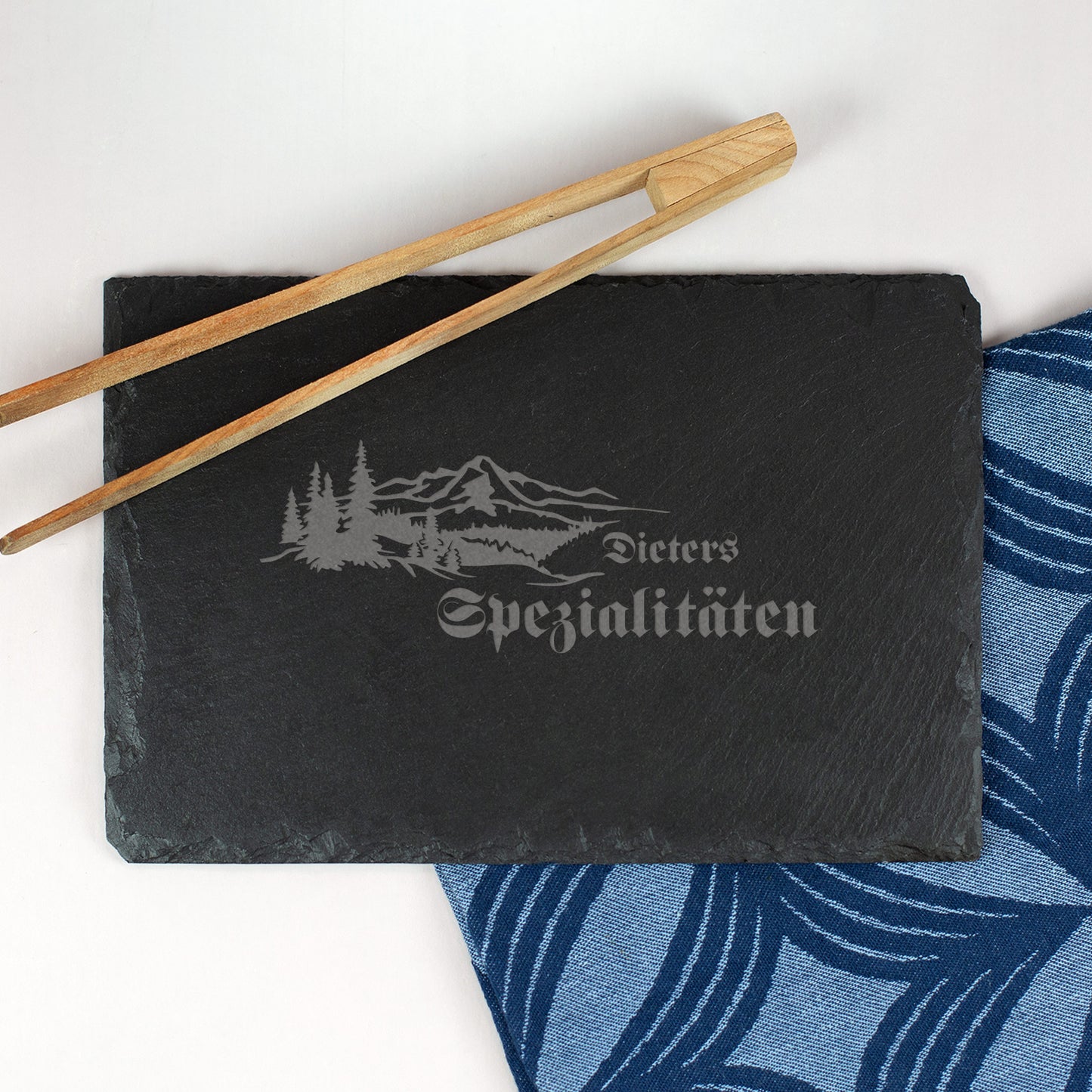 Personalisierte Schieferplatte 30x20 cm mit Gravur „Dieters Spezialitäten“ und Wald-Berge-Motiv, daneben Essstäbchen und Stoffserviette.