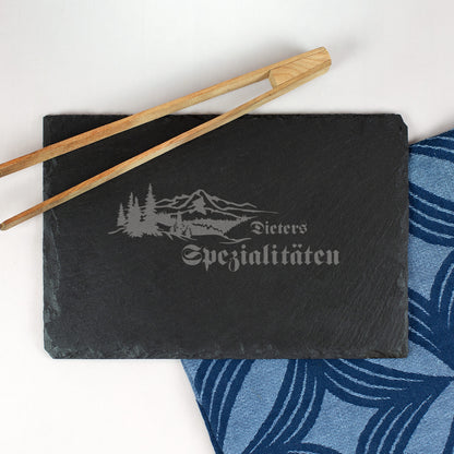 Personalisierte Schieferplatte 30x20 cm mit Gravur „Dieters Spezialitäten“ und Wald-Berge-Motiv, daneben Essstäbchen und Stoffserviette.